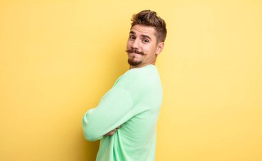 Genç yakışıklı adam neşeyle gülümsüyor, mutlu, tatmin olmuş ve rahatlamış hissediyor, kollarını kavuşturup yana bakıyor. Garip bıyık konsepti