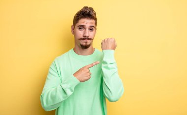 Genç yakışıklı adam sabırsız ve kızgın görünüyor, saati gösteriyor, dakiklik istiyor, zamanında gelmek istiyor. Garip bıyık konsepti