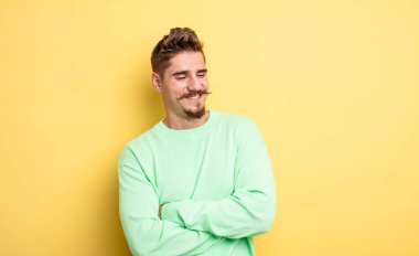 Genç yakışıklı adam kollarını kavuşturup mutlu bir şekilde gülüyor, rahat, pozitif ve tatmin edici bir pozla. Garip bıyık konsepti