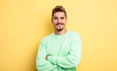 Mutlu, gururlu ve tatmin olmuş kollarını kavuşturarak gülümseyen genç yakışıklı bir adam. Garip bıyık konsepti