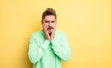 Genç yakışıklı adam şok olmuş ve korkmuş hissediyor, ağzı açık ve elleri yanaklarında korkmuş görünüyor. Garip bıyık konsepti