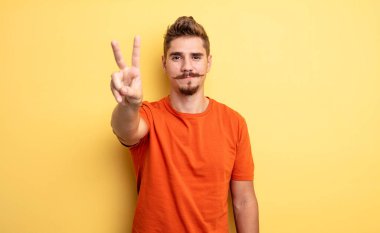 Genç yakışıklı adam gülümsüyor ve arkadaş canlısı görünüyor. İki ya da iki numarayı gösteriyor. Elleri önde, geriye doğru sayıyor. Garip bıyık konsepti