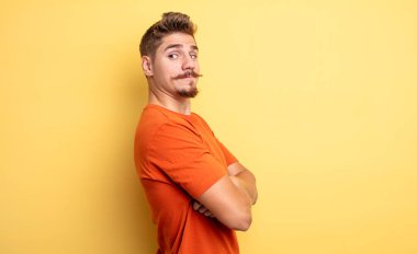 Genç yakışıklı adam neşeyle gülümsüyor, mutlu, tatmin olmuş ve rahatlamış hissediyor, kollarını kavuşturup yana bakıyor. Garip bıyık konsepti