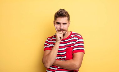 Genç yakışıklı adam ciddi, düşünceli ve güvensiz görünüyor, bir kolu çapraz ve elleri çenesinde, seçenekleri değerlendiriyor. Garip bıyık konsepti