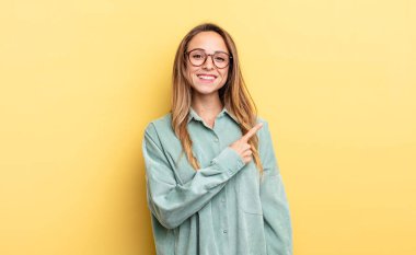 pretty caucasian woman smiling cheerfully, feeling happy and pointing to the side and upwards, showing object in copy space