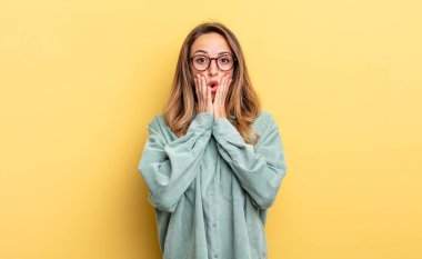 pretty caucasian woman feeling shocked and scared, looking terrified with open mouth and hands on cheeks