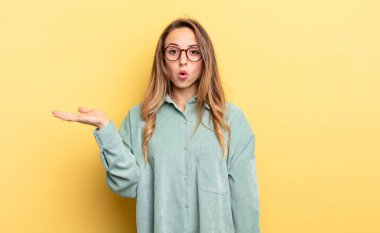 pretty caucasian woman looking surprised and shocked, with jaw dropped holding an object with an open hand on the side