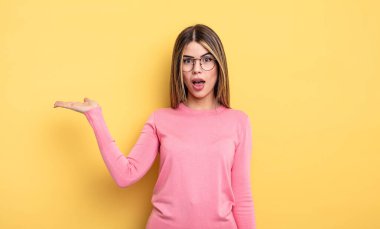 pretty caucasian woman looking surprised and shocked, with jaw dropped holding an object with an open hand on the side