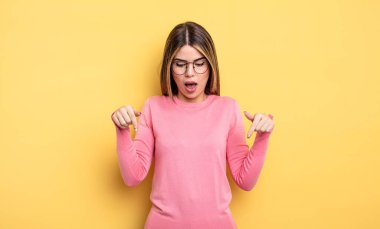 pretty caucasian woman feeling shocked, open-mouthed and amazed, looking and pointing downwards in disbelief and surprise