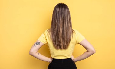 pretty caucasian woman feeling confused or full or doubts and questions, wondering, with hands on hips, rear view