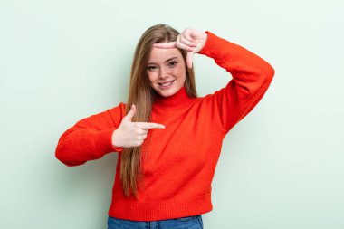 Kafkasyalı kızıl saçlı kadın mutlu, arkadaş canlısı ve pozitif hissediyor, gülümsüyor ve elleriyle bir portre veya fotoğraf çerçevesi yapıyor.