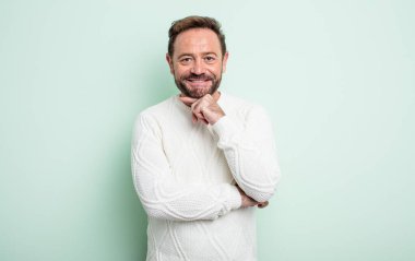 Orta yaşlı yakışıklı adam mutlu ve gülümsüyor. Elleri çenesinde, merak ediyor ya da soru soruyor. Seçenekleri karşılaştırıyor.