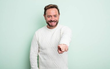 Orta yaşlı yakışıklı bir adam kameraya bakıyor, kendinden emin, arkadaş canlısı bir gülümsemeyle, seni seçiyor.