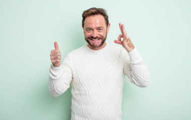 Orta yaşlı yakışıklı bir adam mutlu, şaşkın, tatmin olmuş ve şaşırmış hissediyor, tamam gösteriyor ve el kol hareketi yapıyor, gülümsüyor.