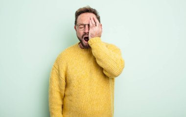 Orta yaşlı yakışıklı bir adam uykulu, sıkılmış ve esniyor, baş ağrısı ve yüzünün yarısını kaplayan bir elle.