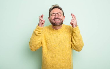 Orta yaşlı yakışıklı bir adam gülümsüyor ve endişeyle iki parmağını çaprazlıyor, endişeli ve iyi şanslar diliyor.