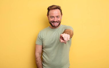 Orta yaşlı yakışıklı bir adam kameraya bakıyor, kendinden emin, arkadaş canlısı bir gülümsemeyle, seni seçiyor.