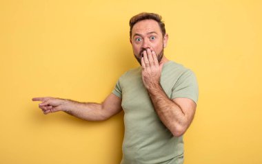 Orta yaşlı yakışıklı bir adam kendini mutlu, şaşkın ve şaşırmış hissediyor. Ağzını eliyle kapatıyor ve yanal fotokopi alanını işaret ediyor.