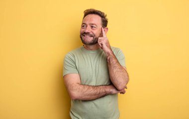 Orta yaşlı yakışıklı adam mutlu bir şekilde gülümsüyor ve hayal kuruyor ya da şüphe ediyor, yan tarafa bakıyor.