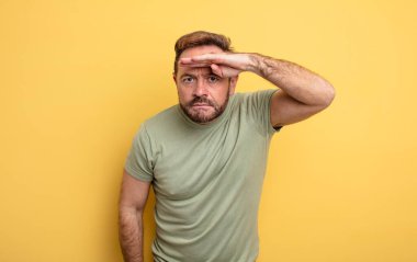 Orta yaşlı yakışıklı adam şaşkın ve şaşkın görünüyor, el üstünde uzak bakıyor, izliyor ya da arıyor