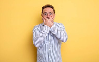 Orta yaşlı yakışıklı bir adam elleri ile ağzını kapatıyor, şok olmuş, şaşırmış bir ifadeyle, bir sır saklıyor ya da 