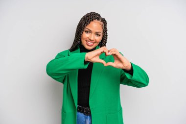 Siyah afro kadın gülümsüyor ve mutlu, sevimli, romantik ve aşık, iki eliyle kalbi şekillendiriyor.