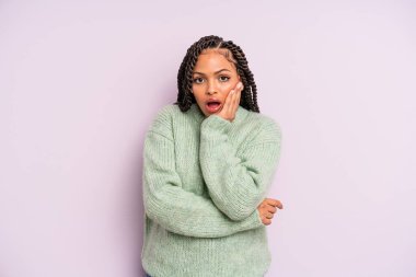 Siyah afro kadın şok ve inançsızlık içinde, elleri yanak ve kolları çapraz, sersemlemiş ve şaşkın hissediyor