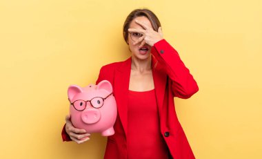businesswoman looking shocked, scared or terrified, covering face with hand. piggy bank concept