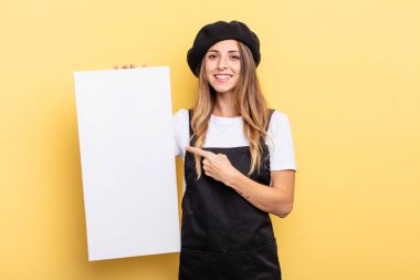 woman artist smiling cheerfully, feeling happy and pointing to the side empty canvas concept