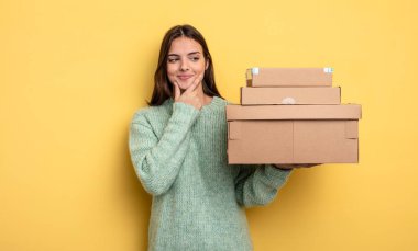 pretty woman smiling with a happy, confident expression with hand on chin packages boxes concept