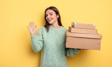 pretty woman smiling happily, waving hand, welcoming and greeting you packages boxes concept