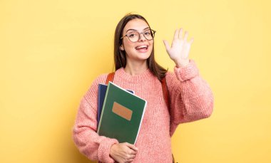 pretty student woman smiling happily, waving hand, welcoming and greeting you