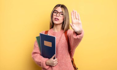Güzel bayan öğrenci ciddi görünüyor. Avuç içi açık duruyor. Durdurma hareketi yapıyor. kitaplar ve sırt çantası