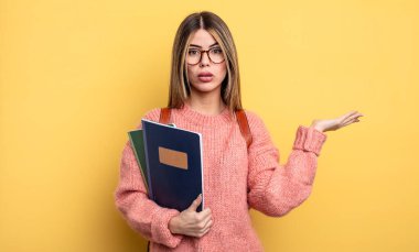 Güzel bir öğrenci kadın şaşırmış ve şok olmuş görünüyor. Çenesi düşmüş ve elinde bir nesne var. kitaplar ve sırt çantası