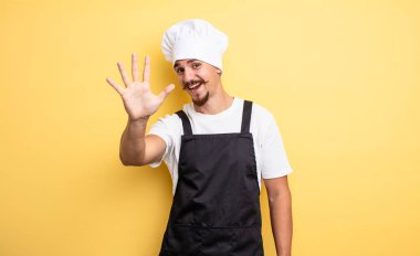 Şef gülümsüyor ve arkadaş canlısı görünüyor, 5 numarayı gösteriyor.