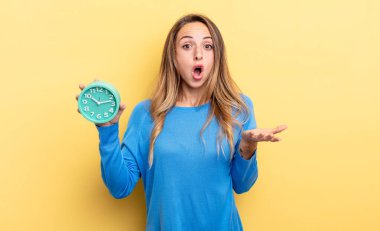 pretty woman amazed, shocked and astonished with an unbelievable surprise holding an alarm clock
