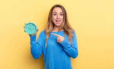 pretty woman looking excited and surprised pointing to the side holding an alarm clock