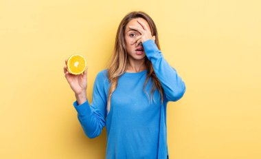 pretty woman looking shocked, scared or terrified, covering face with hand holding half orange