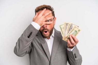 middle age man looking shocked, scared or terrified, covering face with hand. dollar banknotes concept