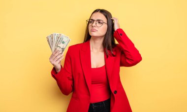 pretty woman feeling puzzled and confused, scratching head. dollar banknotes concept