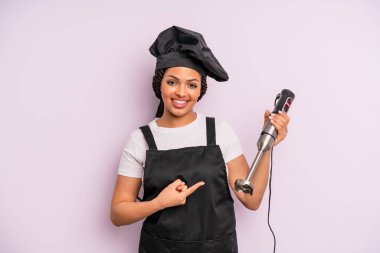 afro black woman smiling cheerfully, feeling happy and pointing to the side. chef with hand blender