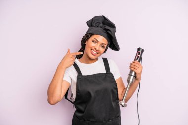 afro black woman smiling confidently pointing to own broad smile. chef with hand blender