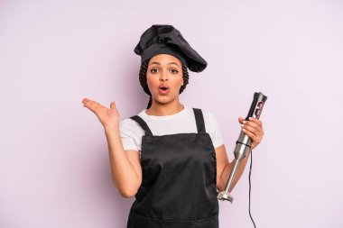 afro black woman looking surprised and shocked, with jaw dropped holding an object. chef with hand blender