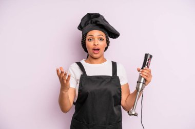 afro black woman feeling extremely shocked and surprised. chef with hand blender