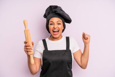 afro black woman feeling shocked,laughing and celebrating success. chef with a roller pin
