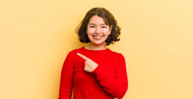 Güzel İspanyol kadın neşeyle gülümsüyor, mutlu hissediyor ve yan ve yukarı doğru işaret ediyor, fotokopi uzayındaki nesneleri gösteriyor.