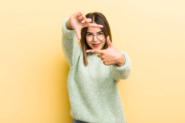 Güzel İspanyol bir kadın mutlu, arkadaş canlısı ve pozitif hissediyor, gülümsüyor ve elleriyle bir portre veya fotoğraf çerçevesi yapıyor.