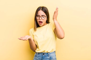 pretty hispanic woman shrugging with a dumb, crazy, confused, puzzled expression, feeling annoyed and clueless