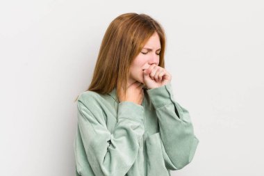 Kızıl saçlı güzel bir kadın boğazı ağrıyan ve grip belirtileri gösteren ağzı kapalı öksüren bir hasta.