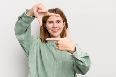 Kızıl saçlı güzel bir kadın mutlu, arkadaş canlısı ve pozitif hissediyor, gülümsüyor ve elleriyle bir portre veya fotoğraf çerçevesi yapıyor.